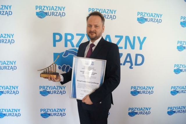Przedstawiciel Urzędu na tle logotypu "Przyjazny Urząd" ze statuetką i dyplomem